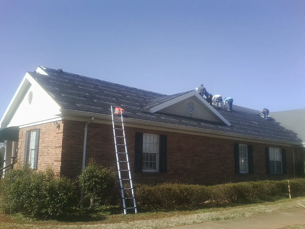 Crew tearing shingles off a brick ranch roof with a ladder set — Emergency Roof Repair work by our crew in Mayville